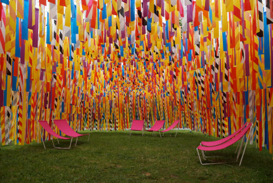 Grassy area with pink lawn chairs topped by a wall of colorful paper strips.